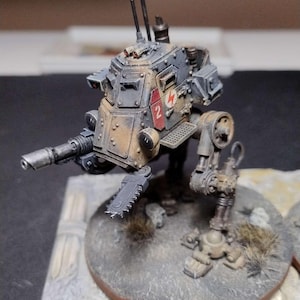 May include: A miniature, detailed model of a war machine, painted in shades of gray, brown, and red. The model features a cannon, saw blade, and a red emblem with the number 2. The model is mounted on a circular base.