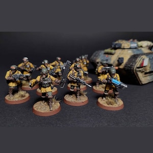Puede incluir: Figuras en miniatura de soldados con uniformes amarillos, cascos y gafas, armados con rifles y armas futuristas. Un tanque de camuflaje está en el fondo. Las figuras están sobre bases marrones, lo que sugiere un juego de mesa o un hobby.