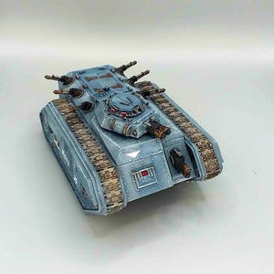Pode incluir: Modelo detalhado de um tanque azul claro com múltiplas torres de canhão e esteiras marrons. O tanque tem um corpo retangular com rebites e uma escotilha na parte superior. O modelo é pintado com efeitos de desgaste.
