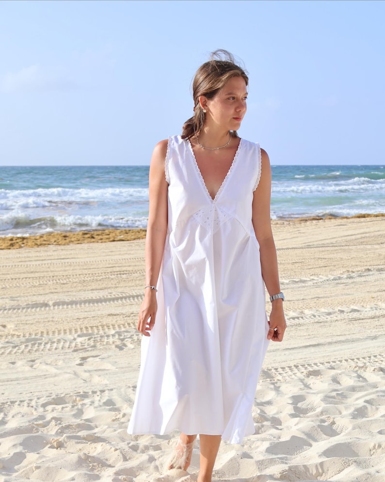 Peut inclure: Une robe midi blanche sans manches avec un d&eacute;collet&eacute; en V profond orn&eacute; d'une fine bordure en dentelle et un d&eacute;tail floral brod&eacute; au centre du corsage. La robe pr&eacute;sente une silhouette d&eacute;contract&eacute;e et fluide qui tombe sous le genou.