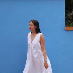 Peut inclure: Une robe midi blanche sans manches dot&eacute;e d'un d&eacute;collet&eacute; en V profond avec une d&eacute;licate bordure en dentelle le long des bords. Le v&ecirc;tement pr&eacute;sente une silhouette ample et fluide avec du tissu fronc&eacute; &agrave; la taille, tombant jusqu'au mollet.