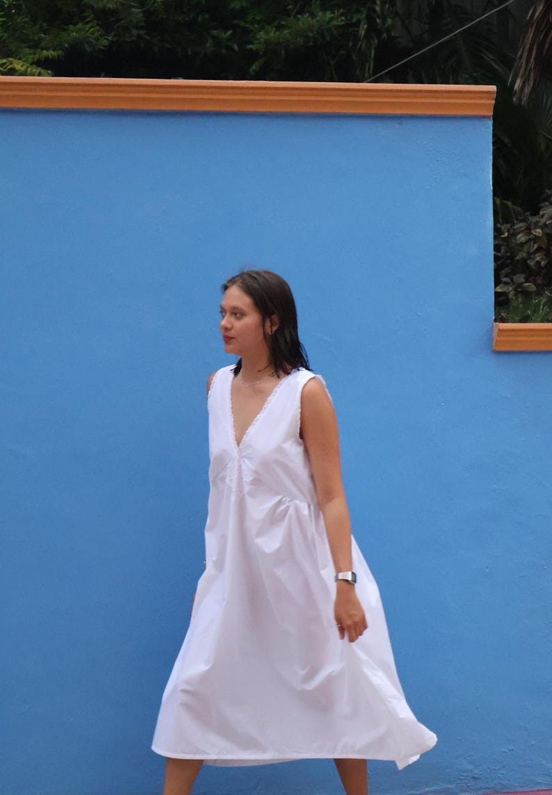 Peut inclure: Une robe midi blanche sans manches dot&eacute;e d'un d&eacute;collet&eacute; en V profond avec une d&eacute;licate bordure en dentelle le long des bords. Le v&ecirc;tement pr&eacute;sente une silhouette ample et fluide avec du tissu fronc&eacute; &agrave; la taille, tombant jusqu'au mollet.