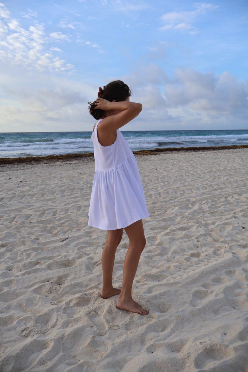 Peut inclure: Une robe courte blanche sans manches avec une taille fronc&eacute;e et une jupe &eacute;vas&eacute;e. Le v&ecirc;tement pr&eacute;sente une encolure ronde simple et une confection en tissu blanc uni, pr&eacute;sent&eacute;e de profil dans un environnement naturel en ext&eacute;rieur.