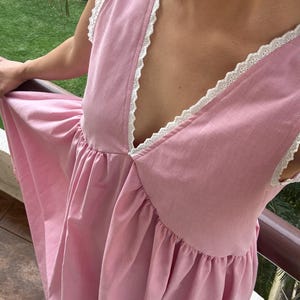 May include: A sleeveless pink dress featuring a deep V-neckline and armholes trimmed with white eyelet lace. The bodice is fitted, transitioning into a gathered, flowing skirt that creates a soft, voluminous silhouette with a solid pastel color.
