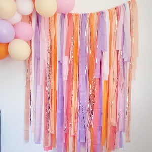 Op de afbeelding: Een kleurrijke feestachtergrond met pastelkleurige stoffen slingers in roze, perzik, oranje en paars. Metallic gouden slingers zijn ertussenin verwerkt. Ballonnen in bijpassende kleuren zijn links gegroepeerd.