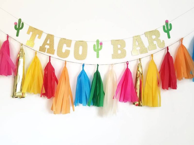 Puede incluir: Un banner de purpurina dorada "Taco Bar" con decoraciones de cactus verdes y borlas de papel de seda de colores.