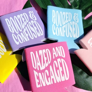 May include: Five colorful can coolers with different humorous phrases printed on them. The phrases are "Boozed & Confused", "Dazed and Engaged", and "Boozed & Confused". The coolers are on a pink surface with a green leaf in the background.