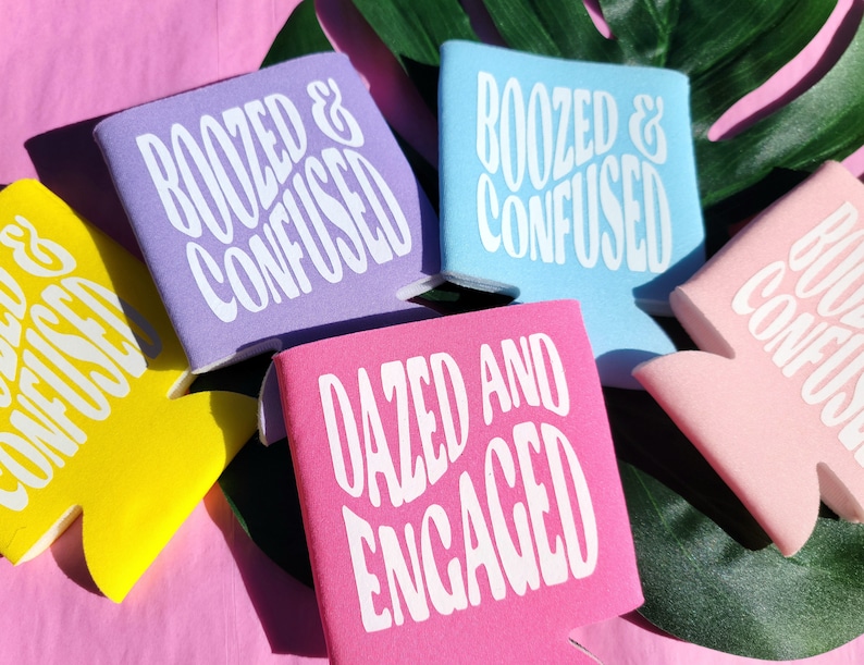 May include: Five colorful can coolers with different humorous phrases printed on them. The phrases are "Boozed & Confused", "Dazed and Engaged", and "Boozed & Confused". The coolers are on a pink surface with a green leaf in the background.