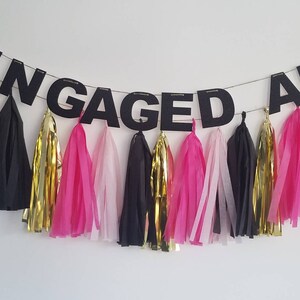 May include: A black banner with the text "ENGAGED AF" hanging from a string with pink, black, and gold tissue paper tassels.