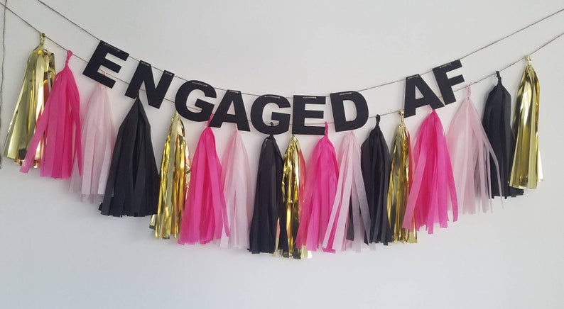 May include: A black banner with the text "ENGAGED AF" hanging from a string with pink, black, and gold tissue paper tassels.