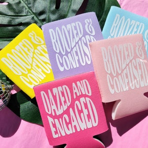 May include: Four colorful can coolers with different phrases printed on them. The phrases are "Roozed & Confused", "Roozed & Confused", "Roozed & Confused", and "Dazed and Engaged".