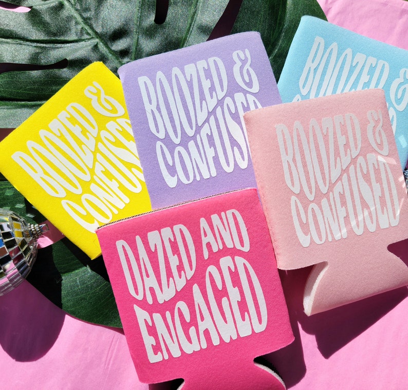 May include: Four colorful can coolers with different phrases printed on them. The phrases are "Roozed & Confused", "Roozed & Confused", "Roozed & Confused", and "Dazed and Engaged".