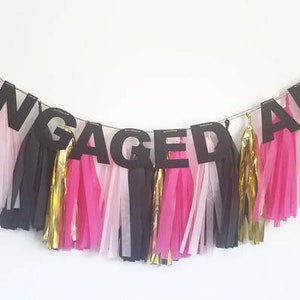May include: Black paper banner with the word "ENGAGED AF" in large letters. The banner is decorated with pink, white, and gold tissue paper tassels.