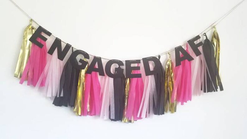 May include: Black paper banner with the word "ENGAGED AF" in large letters. The banner is decorated with pink, white, and gold tissue paper tassels.