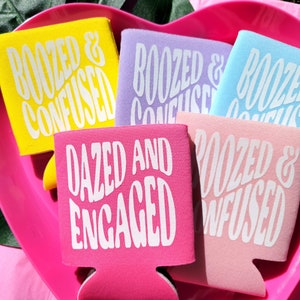 May include: Five colorful can coolers with different humorous phrases printed on them. The phrases are "Roozed & Confused", "Dazed and Engaged", and "Roozed & Confused" in different colors.