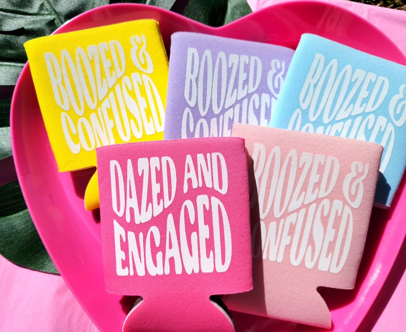 May include: Five colorful can coolers with different humorous phrases printed on them. The phrases are "Roozed & Confused", "Dazed and Engaged", and "Roozed & Confused" in different colors.