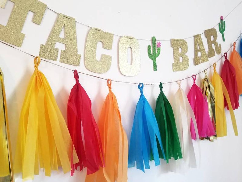 Puede incluir: Una pancarta de brillantina dorada con las palabras "Taco Bar" y un cactus con flores rosas. La pancarta est&aacute; adornada con borlas de papel de seda de colores en amarillo, rojo, naranja, azul, verde, blanco y dorado.