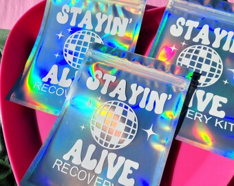 May include: Three iridescent holographic zippered pouches with the text "Stayin' Alive Recovery Kit" and a disco ball graphic.