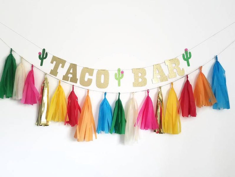 Puede incluir: Un banner de purpurina dorada "Taco Bar" con decoraciones de cactus verdes y borlas de papel de seda de colores.