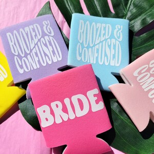 May include: Set of six colorful can coolers with different phrases. The coolers are purple, light blue, pink, and white. The phrases on the coolers are "Boozed & Confused", "Bride", and "Boozed & Confused".