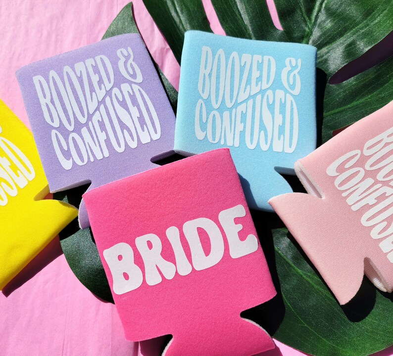 May include: Set of six colorful can coolers with different phrases. The coolers are purple, light blue, pink, and white. The phrases on the coolers are "Boozed & Confused", "Bride", and "Boozed & Confused".