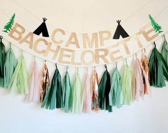 May include: A gold glitter banner with the text "CAMP BACHELORETTE" and black paper tent decorations. Green and pink tissue paper tassels hang below the banner.