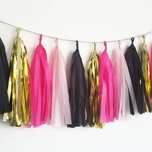 May include: A garland of tissue paper tassels in pink, black, and gold. The tassels are strung together with a thin string and are hanging from a white wall.