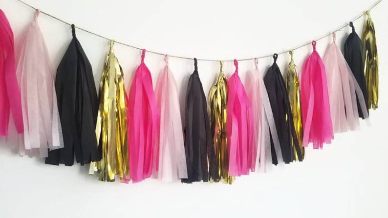 May include: A garland of tissue paper tassels in pink, black, and gold. The tassels are strung together with a thin string and are hanging from a white wall.