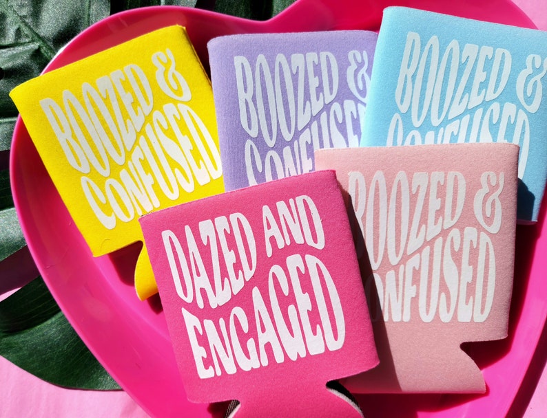 May include: A set of six colorful can coolers with white text. The coolers feature the phrases "Boozed & Confused" and "Dazed and Engaged".
