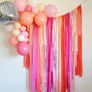 Op de afbeelding: Een feestelijke achtergrond met een cluster van roze, perzik en witte ballonnen, waaronder een discobal. De achtergrond bestaat uit kleurrijke, verticale linten in tinten roze, oranje en zilver, wat een levendige en feestelijke sfeer cre&euml;ert.
