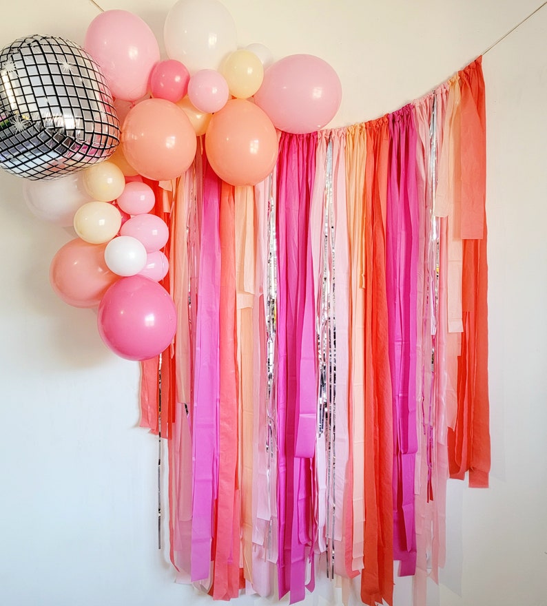 Op de afbeelding: Een feestelijke achtergrond met een cluster van roze, perzik en witte ballonnen, waaronder een discobal. De achtergrond bestaat uit kleurrijke, verticale linten in tinten roze, oranje en zilver, wat een levendige en feestelijke sfeer cre&euml;ert.