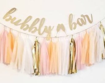 May include: Gold "bubbly bar" sign with a string of pink, peach, white, and gold tissue paper tassels.