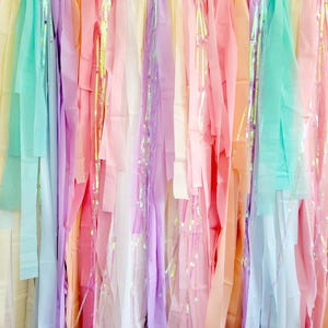 Op de afbeelding: Een kleurrijke achtergrond van hangende linten in pastelkleuren van roze, paars, geel en blauw. Sommige linten hebben iriserende accenten, waardoor een glinsterend effect ontstaat. De linten zijn verticaal gerangschikt, waardoor een textuur en visueel aantrekkelijke weergave ontstaat.