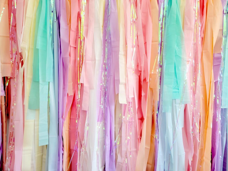 Op de afbeelding: Een kleurrijke achtergrond van hangende linten in pastelkleuren van roze, paars, geel en blauw. Sommige linten hebben iriserende accenten, waardoor een glinsterend effect ontstaat. De linten zijn verticaal gerangschikt, waardoor een textuur en visueel aantrekkelijke weergave ontstaat.