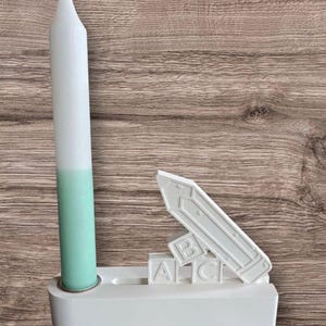 May include: A white and mint green candle stands in a white holder, next to a white pencil-shaped object and ABC blocks. The candle has a two-tone design. The objects are on a wooden surface.