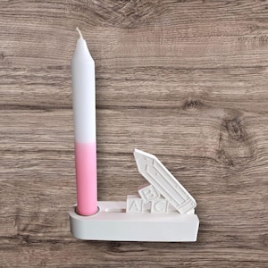 May include: A white and pink candle in a white holder with a pencil and ABC blocks design. The candle is white at the top and fades to pink at the bottom. The holder is on a wooden surface.