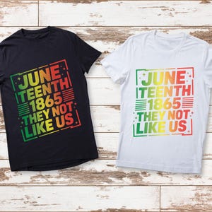 Pode incluir: Duas camisetas, uma preta e outra branca, com o texto "June 1865 They Not Like Us" em um gradiente de verde, amarelo e vermelho. As camisetas são de manga curta e têm gola redonda.