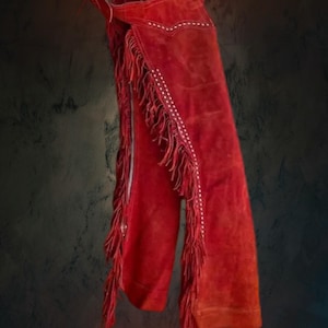 May include: Red suede chaps with white stitching and fringe detailing. The chaps are hanging against a dark background. The chaps are a western wear item.