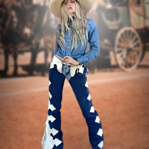 May include: A person wearing a denim shirt, blue pants with white star-shaped accents, and a cream-colored cowboy hat. The outfit has a Western theme, with a blurred background of a horse-drawn carriage.