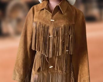 May include: A brown suede jacket with long fringe detailing and a matching skirt. The jacket has a collared neckline and button closures. A white cowboy hat with a turquoise band sits atop the mannequin's head. The outfit is a vintage western style.