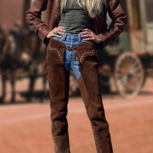May include: A brown leather jacket and chaps are worn over a green top and blue jeans. The chaps have decorative stitching. The outfit has a Western theme.