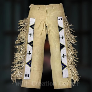 Puede incluir: Pantalones de ante color beige con flecos decorativos a lo largo de las costuras exteriores. Los pantalones presentan un diseño de cuentas blancas y azules con cruces negras y triángulos azules. Un artículo clásico del oeste.