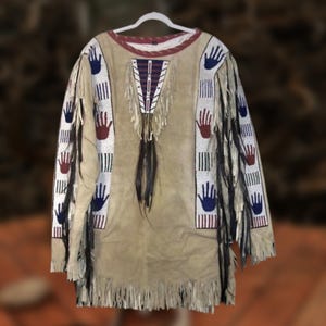 Op de afbeelding: Een bruin overhemd in Native American-stijl met handgeschilderde blauwe en rode handafdrukken op de mouwen. Het overhemd heeft franjes langs de onderkant en mouwen, met een kralenontwerp op de voorkant en mouwen. De kraag is rood afgewerkt.