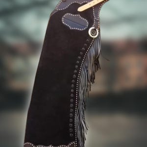 May include: Black leather chaps with white stitching and fringe. The chaps have a decorative top with a leather strap and buckle. The chaps are designed for riding and have a classic Western style.