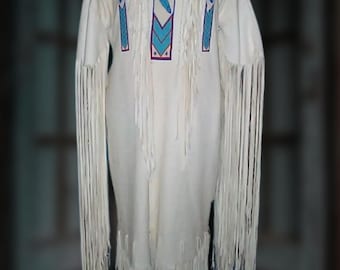 May include: Cream-coloured dress with long sleeves and fringe detailing. The dress features turquoise and purple geometric patterns on the bodice and sleeves. The fringe hangs from the sleeves and the bottom hem. The dress is displayed on a mannequin.