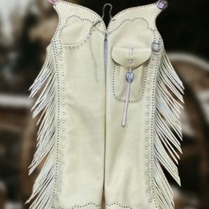 May include: Cream-colored leather chaps with decorative stitching and fringe. Features include a small pocket, silver-toned hardware, and a decorative top edge. The chaps are designed with a western theme.