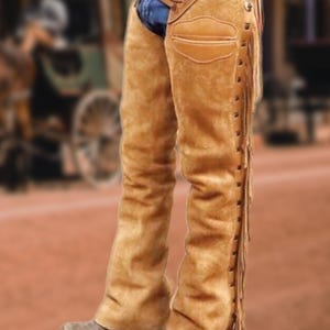 May include: Tan suede leather chaps with fringe detailing along the outer seams. The chaps feature a fitted design with a pocket on the right leg and are worn over blue jeans. The image has a western theme.