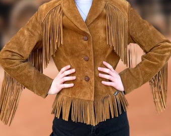 May include: A brown suede jacket with fringe detailing on the sleeves and hem. The jacket has a button-up front and a classic collar. The jacket is a medium brown color and appears to be vintage.