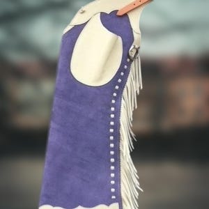 May include: A single purple suede leather chaps with cream-colored accents and fringe. The chaps feature a scalloped bottom edge, a cream-colored pocket, and a leather belt with a silver buckle.
