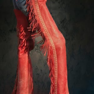 May include: Red leather chaps with fringe detailing and studded accents. The chaps are worn over blue denim jeans. The chaps have a western style and are suitable for a variety of occasions.
